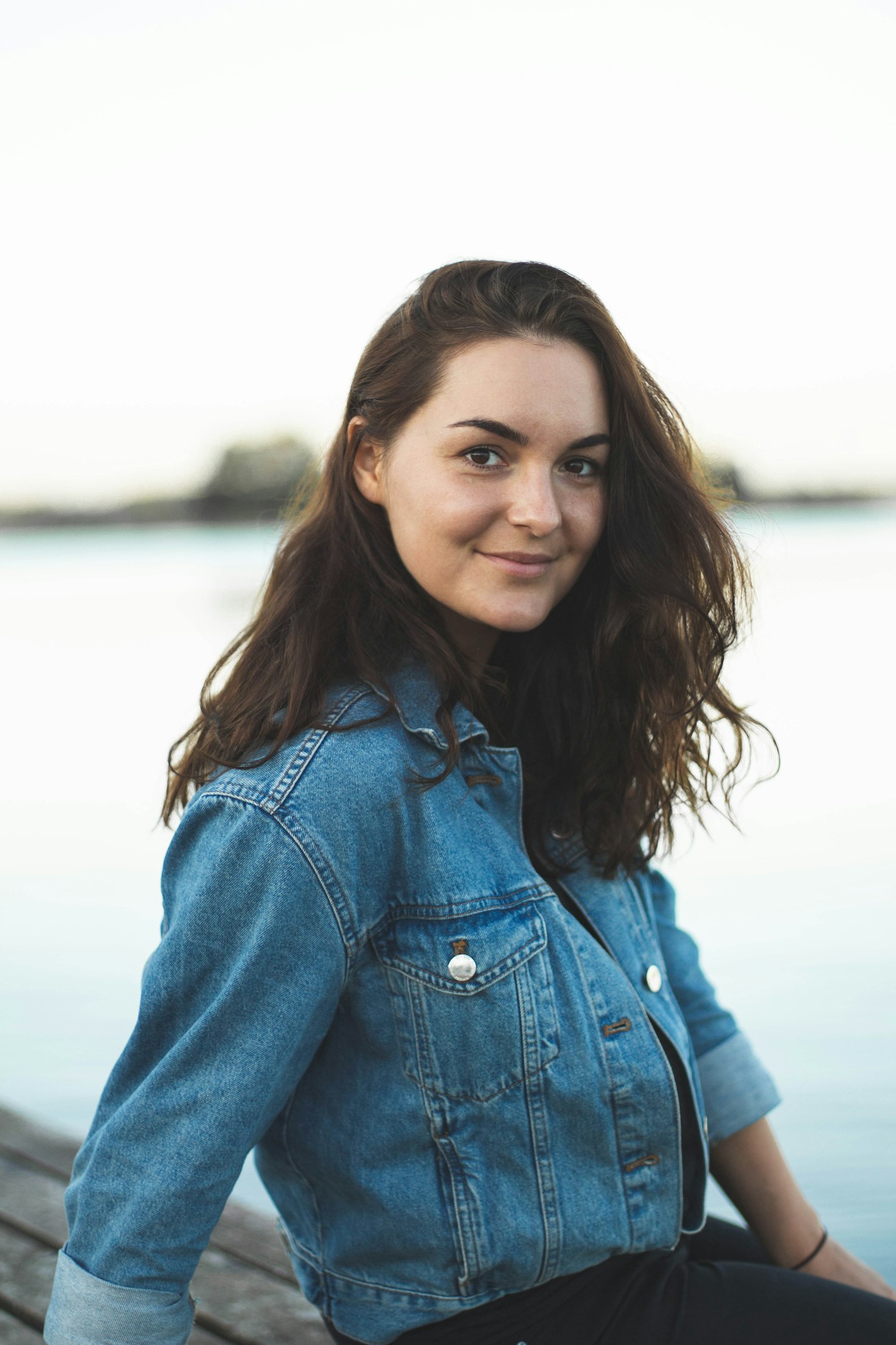 Smiling woman in a denim jacket seated by a calm lake